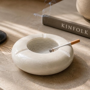May include: A circular white marble ashtray with a lit cigarette. The ashtray has a central indentation. A glass of amber liquid and a book titled "KINFOLK" are also visible in the image.