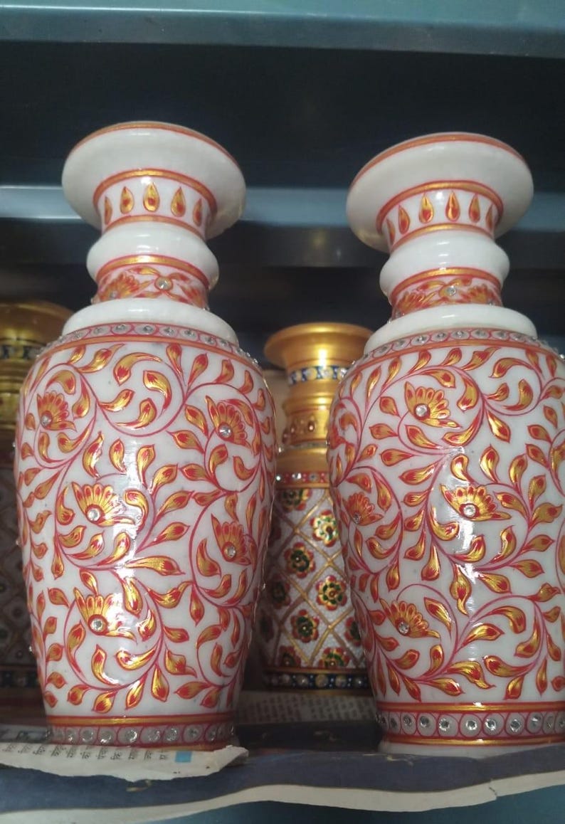 K&ouml;nnte beinhalten: Zwei Vasen aus wei&szlig;em Marmor mit handgemalten, gold-roten floralen Rankenmustern. Jede Vase hat einen ausgestellten Rand, einen bauchigen K&ouml;rper und ist mit kleinen, klaren eingelegten Steinen am Hals und am Fu&szlig; verziert.