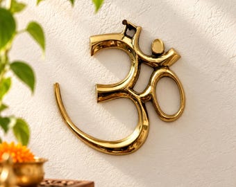 May include: A polished gold-toned metal wall hanging featuring the Om symbol. The design consists of a curved base, a central loop, and a top stroke with a small dot, all rendered in a smooth, reflective finish with clean, rounded edges.