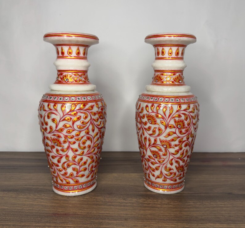 K&ouml;nnte beinhalten: Ein Paar Vasen aus wei&szlig;em Marmor mit handgemalten roten und goldenen Blumenrankenmustern. Die Vasen sind mit kleinen eingelegten Kristallen am Hals und K&ouml;rper verziert und weisen einen ausgestellten Rand sowie eine konische Form auf.