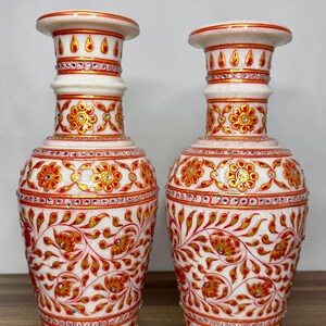K&ouml;nnte beinhalten: Zwei Vasen aus wei&szlig;em Marmor mit handgemalten Blumenmustern in Rot und Gold. Die Vasen sind mit kleinen eingebetteten Kristallen verziert und verf&uuml;gen &uuml;ber einen ausgestellten Rand, einen schlanken Hals und einen bauchigen K&ouml;rper.
