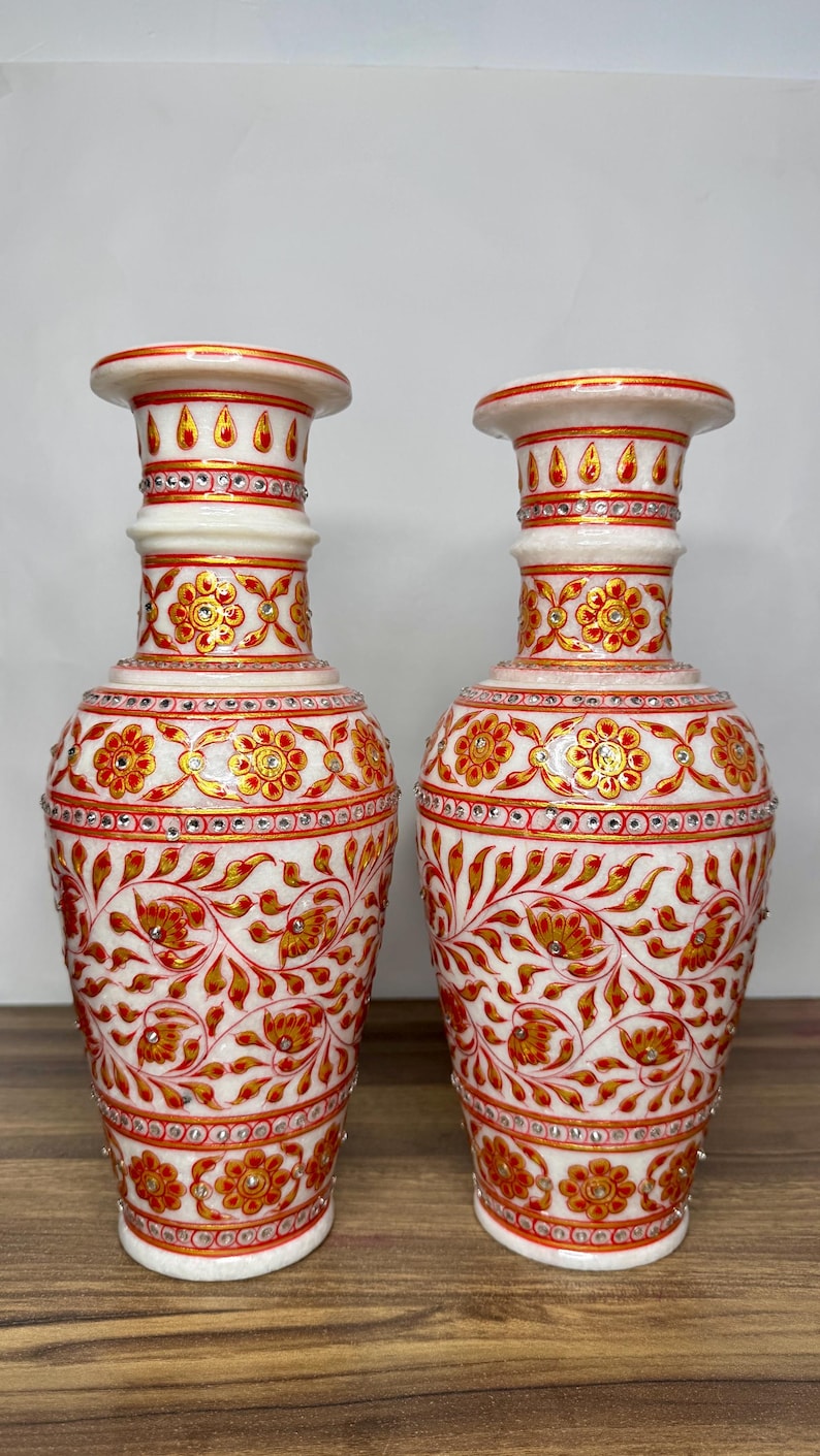 K&ouml;nnte beinhalten: Zwei Vasen aus wei&szlig;em Marmor mit handgemalten Blumenmustern in Rot und Gold. Die Vasen sind mit kleinen eingebetteten Kristallen verziert und verf&uuml;gen &uuml;ber einen ausgestellten Rand, einen schlanken Hals und einen bauchigen K&ouml;rper.