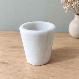 May include: A white marble mortar with a smooth, tapered exterior and a wide, circular opening. The stone features subtle grey veining throughout its surface, showcasing a solid, heavy construction with a thick rim and a flat, stable base.