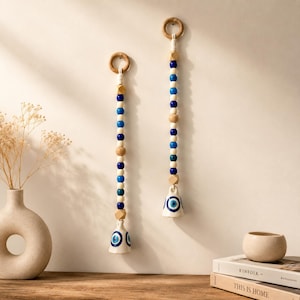 May include: Two vertical wall hangings featuring strings of blue, white, and natural wood beads suspended from wooden rings. Each string terminates in a white ceramic bell decorated with a blue and white evil eye symbol painted on the surface.