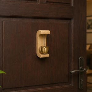Può includere: Un campanello da porta con un supporto in legno chiaro a forma di C fissato su una porta marrone scuro. Una piccola campana in ottone lucido pende dal centro del telaio in legno tramite un sottile cordino nero sopra una sfera percussiva.