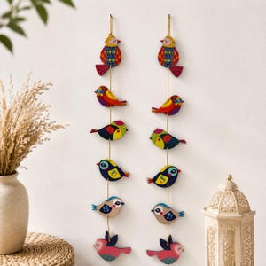 Puede incluir: Dos adornos de pared verticales que presentan una serie de seis siluetas de pájaros de madera coloridos suspendidos en finos hilos dorados. Cada pájaro muestra un patrón y una combinación de colores únicos, incluyendo tonos rojos, azules, amarillos y verdes.