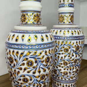 K&ouml;nnte beinhalten: Zwei Vasen aus wei&szlig;em Marmor mit handgemalten Blumenmustern in Gold und Blau. Die Vasen sind mit eingelassenen, klaren Kristallen verziert, die in horizontalen B&auml;ndern und in den Mitten der gemalten Bl&uuml;ten angeordnet sind.