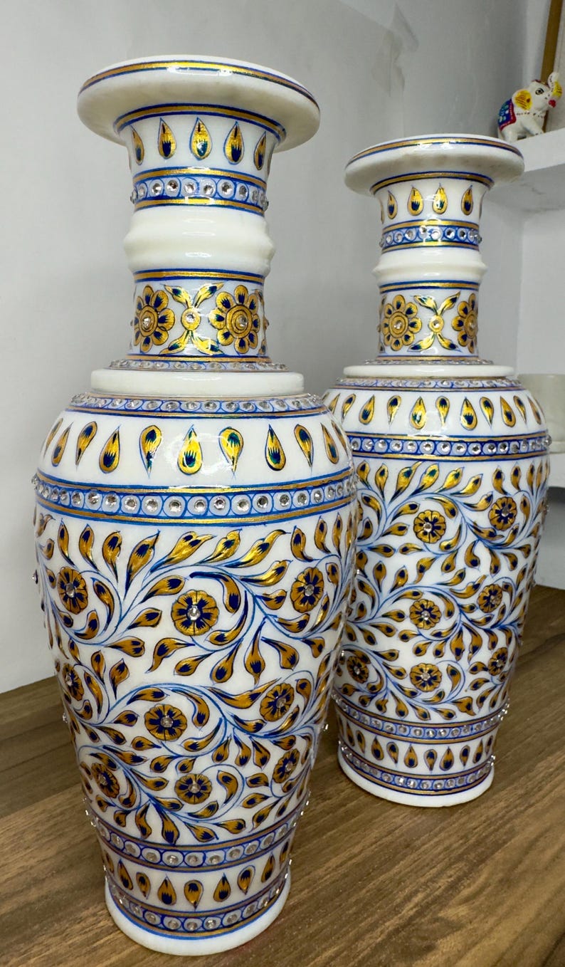 K&ouml;nnte beinhalten: Zwei Vasen aus wei&szlig;em Marmor mit handgemalten Blumenmustern in Gold und Blau. Die Vasen sind mit eingelassenen, klaren Kristallen verziert, die in horizontalen B&auml;ndern und in den Mitten der gemalten Bl&uuml;ten angeordnet sind.