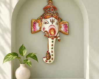 May include: A wall-mounted decorative mask of the deity Ganesha, featuring a white base with intricate hand-painted details in red, pink, green, and gold. The design includes a crown, large ears with pink accents, and a trunk with colourful patterns.