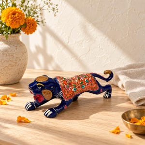 May include: A hand-painted blue tiger figurine in a pouncing pose, featuring a vibrant orange saddle blanket decorated with intricate floral patterns in green, pink, and white, with gold-toned accents on the head, paws, and tail tip.