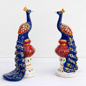 Könnte beinhalten: Zwei handbemalte Pfauenfiguren mit tiefblauen Körpern und goldenen Kämmen. Jeder Vogel steht auf einem rot-weißen Sockel, der mit grünen und roten Punkten verziert ist. Die Flügel und Schwänze zeigen komplizierte Blumenmuster in Rot, Grün und Gelb.