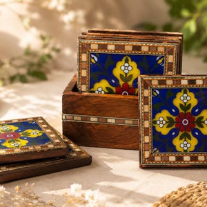 May include: A set of square wooden coasters with a floral design. The coasters have a blue background with yellow and white floral patterns, framed by a mosaic border. They are presented in a wooden box.