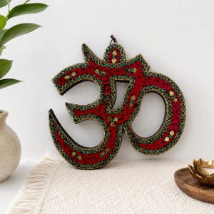 Könnte beinhalten: Ein Om-Symbol als Wandbehang mit einem Mosaikdesign aus roten Glasstücken, akzentuiert durch kleine grüne runde Perlen und goldfarbene Metalleinlagen. Die gesamte Struktur ist mit einem dunklen Rand und einer kleinen Aufhängeschlaufe versehen.