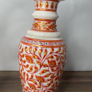 K&ouml;nnte beinhalten: Eine Vase aus wei&szlig;em Marmor mit handgemalten roten und goldenen Blumenmustern. Die Vase ist mit eingelegten klaren Kristallen verziert, die in horizontalen B&auml;ndern um den Hals und den K&ouml;rper angeordnet sind, mit einem ausgestellten Rand.