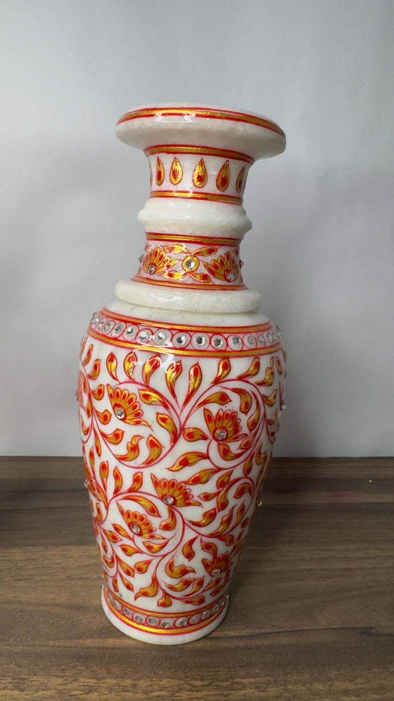 K&ouml;nnte beinhalten: Eine Vase aus wei&szlig;em Marmor mit handgemalten roten und goldenen Blumenmustern. Die Vase ist mit eingelegten klaren Kristallen verziert, die in horizontalen B&auml;ndern um den Hals und den K&ouml;rper angeordnet sind, mit einem ausgestellten Rand.