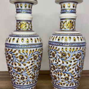 K&ouml;nnte beinhalten: Ein Paar Vasen aus wei&szlig;em Marmor mit handgemalten floralen Mustern in Gold und Blau. Die Vasen haben einen schlanken Hals, einen ausgestellten Rand und einen bauchigen K&ouml;rper, der mit Rankenmotiven, kleinen goldenen Bl&uuml;ten und blauen Akzenten verziert ist.