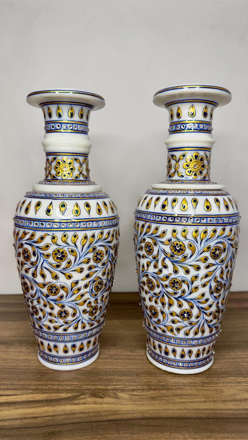 K&ouml;nnte beinhalten: Ein Paar Vasen aus wei&szlig;em Marmor mit handgemalten floralen Mustern in Gold und Blau. Die Vasen haben einen schlanken Hals, einen ausgestellten Rand und einen bauchigen K&ouml;rper, der mit Rankenmotiven, kleinen goldenen Bl&uuml;ten und blauen Akzenten verziert ist.