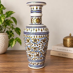 K&ouml;nnte beinhalten: Eine Vase aus wei&szlig;em Marmor mit handgemalten Blumenmustern in Gold und Blau. Die Oberfl&auml;che ist mit kleinen, reflektierenden eingelegten Steinen verziert, die in geometrischen B&auml;ndern und Blattmotiven auf dem Gef&auml;&szlig; angeordnet sind.