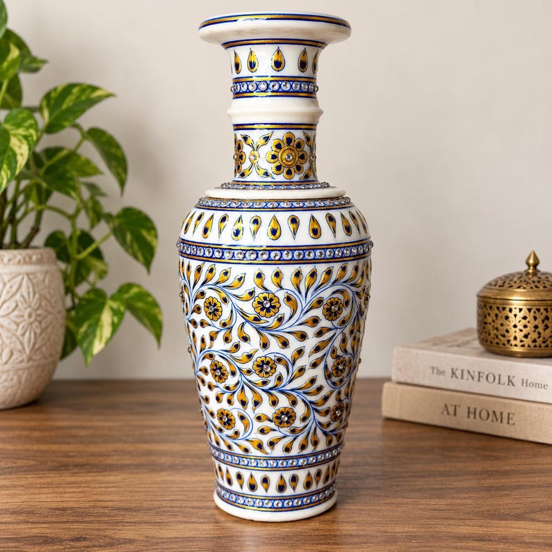 K&ouml;nnte beinhalten: Eine Vase aus wei&szlig;em Marmor mit handgemalten Blumenmustern in Gold und Blau. Die Oberfl&auml;che ist mit kleinen, reflektierenden eingelegten Steinen verziert, die in geometrischen B&auml;ndern und Blattmotiven auf dem Gef&auml;&szlig; angeordnet sind.