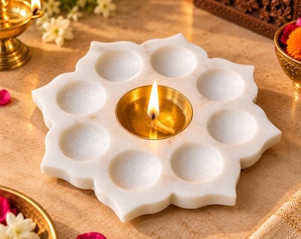 May include: A white marble lotus-shaped candle holder featuring eight circular indentations surrounding a central brass cup. A single lit wick burns within the brass cup, casting a warm glow. The base is carved into a flower petal design.