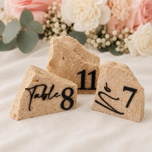 May include: Three beige stone table number holders with black numbers. One reads "Table 8", another "11", and the third "7". The background features a soft white cloth and a bouquet of pink and white flowers.