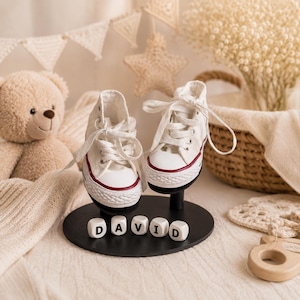 Könnte beinhalten: Ein Paar weiße Baby-Leinenschuhe mit roter Umrandung und weißen Schnürsenkeln, ausgestellt auf einem schwarzen Ständer. Das Wort "DAVID" ist mit weißen Buchstabenblöcken geschrieben. Die Schuhe sind etwa 10 cm lang.