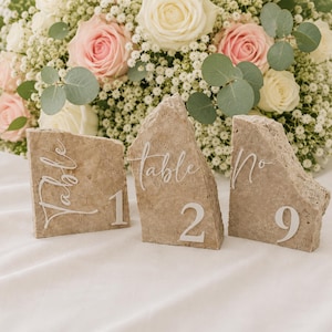 May include: Three stone table number holders with the word "Table" and numbers 1, 2, and 9 in white script. The stone is a light brown color. A bouquet of white and pink roses and greenery is in the background.