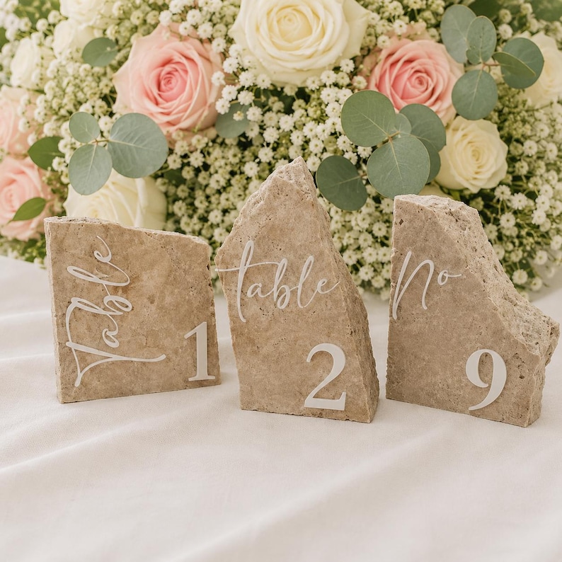 May include: Three stone table number holders with the word "Table" and numbers 1, 2, and 9 in white script. The stone is a light brown color. A bouquet of white and pink roses and greenery is in the background.