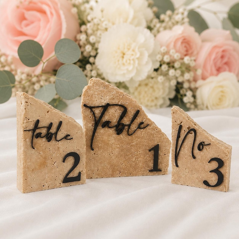 May include: Three beige stone table number holders with black lettering and numbers. The holders are in the shape of a triangle. The text "Table" is on two holders, and "No" is on the third. Numbers 1, 2, and 3 are visible.