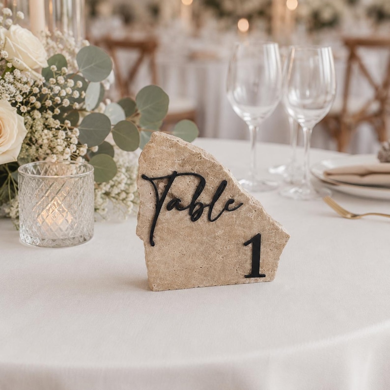 May include: A natural stone table number with the word "Table" in black script and the number "1". The stone is beige and sits on a white tablecloth. The background includes flowers, candles, and glassware.