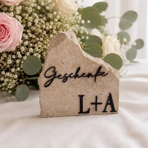 Könnte beinhalten: Ein hellbeiger Stein mit dem Wort "Geschenke" und den Initialen "L+A" in Schwarz. Der Stein ist grob behauen und steht auf einer weißen Oberfläche, mit einem Blumenstrauß im Hintergrund.