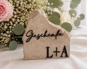 May include: A light beige stone with the word "Geschenke" and the initials "L+A" in black. The stone is rough-hewn and stands on a white surface, with a bouquet of flowers in the background.