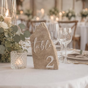 May include: A natural stone table number 2, with the word "table" in cursive script. The stone is beige and stands on a white tablecloth. Wine glasses and a candle holder are in the background, suggesting a wedding or event setting.