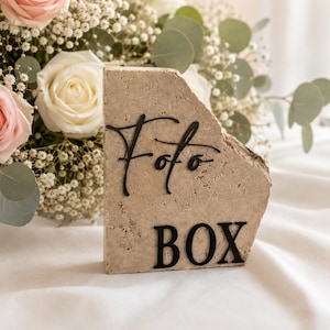 Könnte beinhalten: Ein beigefarbener Steinblock mit den Worten "Foto BOX" in schwarzer Schrift. Der Block hat eine raue, natürliche Textur. Er steht vor einem Hintergrund aus weißem Stoff und einem Strauß Rosen und Grün.