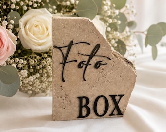 May include: A beige stone block with the words "Foto BOX" in black lettering. The block has a rough, natural texture and is set against a backdrop of white fabric and a bouquet of roses and greenery.