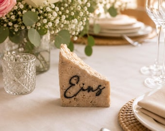 May include: A beige, wedge-shaped place card holder with the word "Eins" in black script. The holder sits on a white tablecloth, with a floral arrangement and glassware in the background. The scene suggests a wedding or formal event.