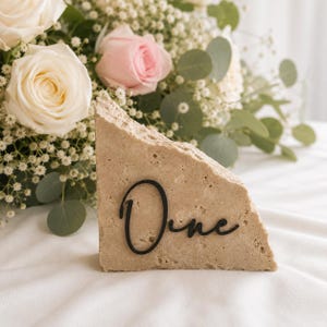 Könnte beinhalten: Ein Tischaufsteller aus beigem Stein mit dem Wort "One" in schwarzer Schrift. Der Stein hat eine raue, strukturierte Oberfläche. Im Hintergrund sind weiße und rosa Rosen und Grün zu sehen.