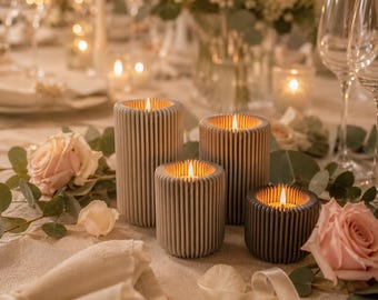 May include: Four lit pillar candles of varying heights, with a ribbed design, sit on a table. The candles are in shades of gray and brown. The scene includes flowers, greenery, and glassware, creating a warm, inviting ambiance.