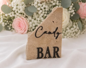 May include: A beige stone candy bar sign with black lettering. The sign is angled and has the words "Candy" and "BAR" in a decorative font. The background features a bouquet of pink and white roses and baby's breath.
