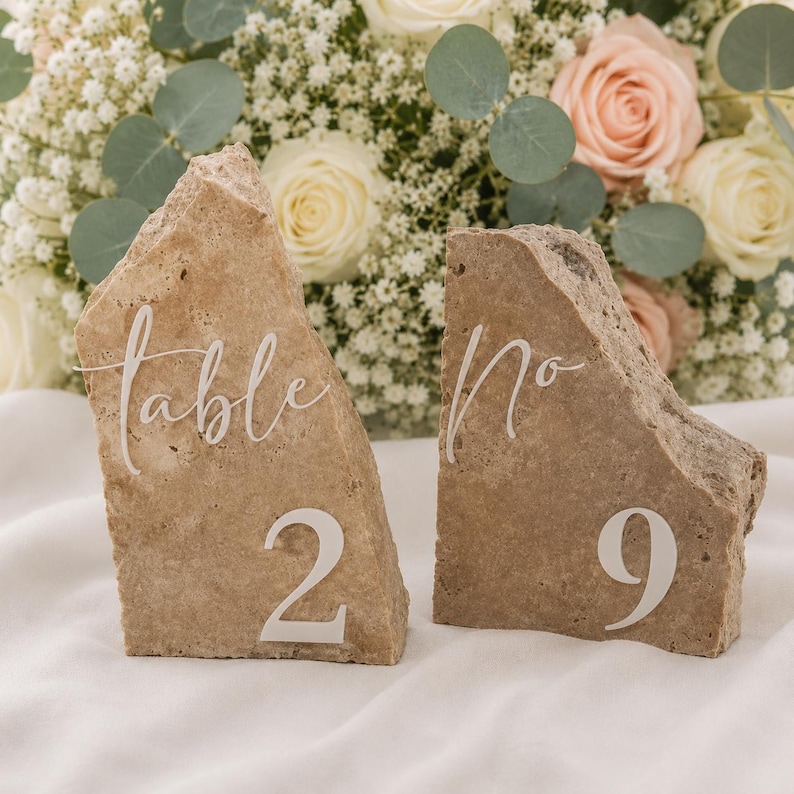 May include: Two natural stone table number signs. One sign reads "table 2" and the other reads "No 9" in white script. The signs are a light brown color and are set against a backdrop of white and pink flowers.