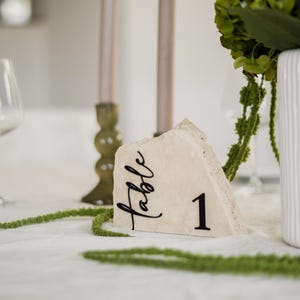 Könnte beinhalten: Ein beigefarbenes Tischnummernschild aus Naturstein mit rauen Kanten und dem schwarzen Aufdruck "table 1" in einer Kombination aus Schreibschrift und serifenloser Schrift. Das Stück hat eine unebene Oberkante und einen flachen Boden.