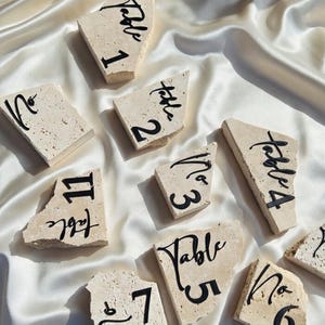 May include: A collection of beige stone table numbers, each with a unique shape and black calligraphy. Numbers 1 through 7 and 11 are visible. The stones rest on a white satin surface, creating an elegant display.