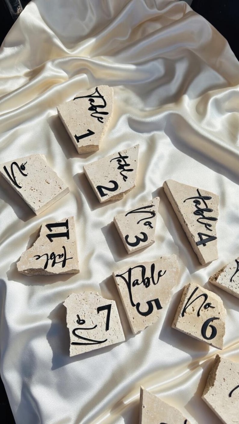 May include: A collection of beige stone table numbers, each with a unique shape and black calligraphy. Numbers 1 through 7 and 11 are visible. The stones rest on a white satin surface, creating an elegant display.