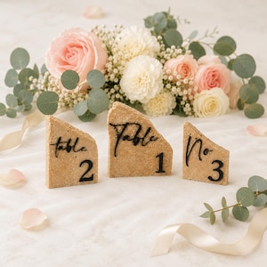 Op de afbeelding: Drie beige stenen tafelnummerhouders met zwarte tekst met de tekst "Tafel 1", "Tafel 2" en "Nr. 3". De houders zijn geplaatst voor een bloemstuk van roze en witte rozen en groen.