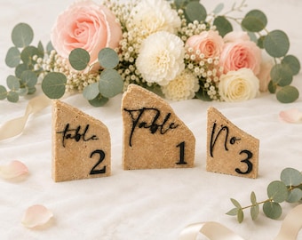 May include: Three beige stone table number holders with black script reading "Table 1", "Table 2", and "No. 3". The holders are arranged in front of a floral arrangement of pink and white roses and greenery.