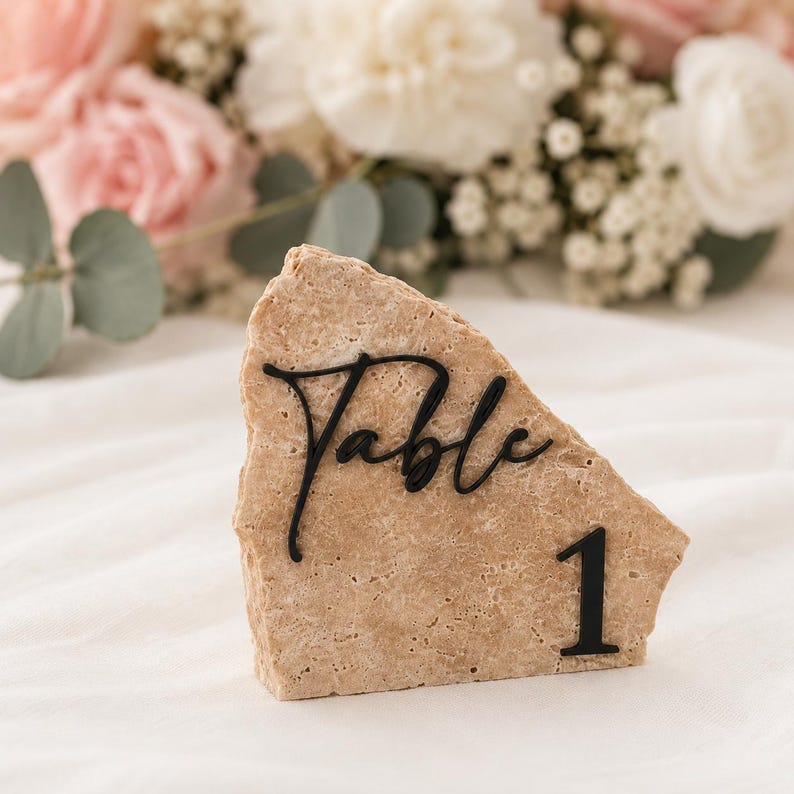 May include: A beige stone table number with the word "Table" in black script and the number "1". The stone has a rough, textured surface. Soft focus background of pink and white flowers.