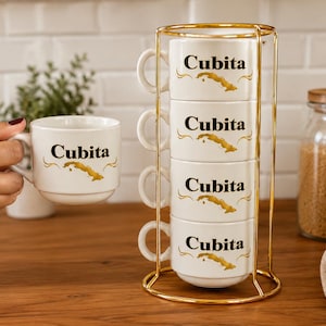 May include: A set of four white ceramic mugs stacked vertically in a gold-toned metal wire rack. Each mug features the black text Cubita above a gold-colored map of Cuba. One matching mug is held separately to the side of the rack.
