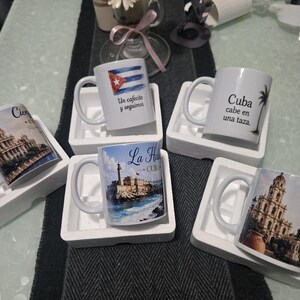 May include: Six white ceramic coffee mugs with Cuban designs. One mug has the Cuban flag and text, another a palm tree and text, and others display architectural images with text. Each mug is in a white foam holder, presented on a table.