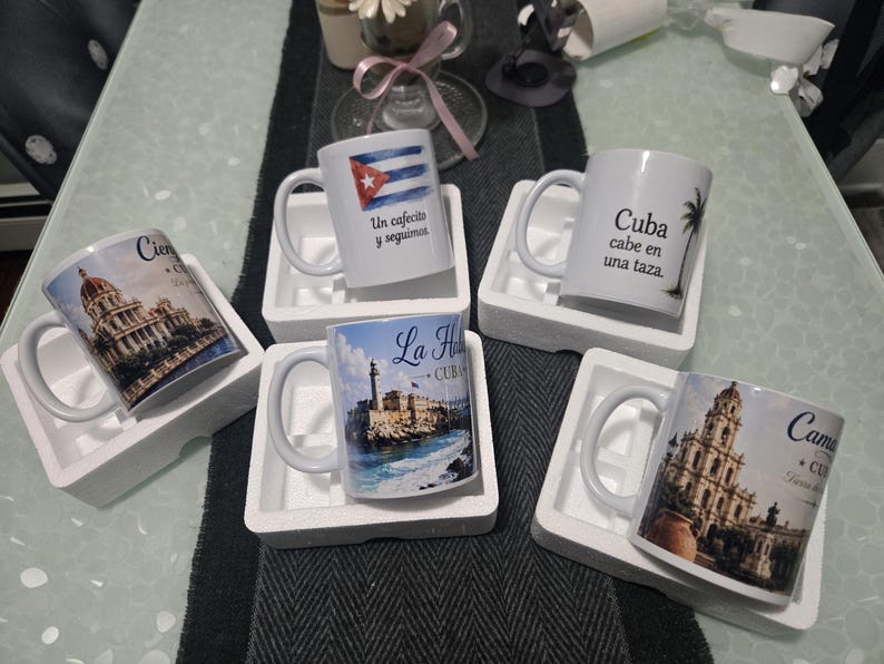 May include: Six white ceramic coffee mugs with Cuban designs. One mug has the Cuban flag and text, another a palm tree and text, and others display architectural images with text. Each mug is in a white foam holder, presented on a table.