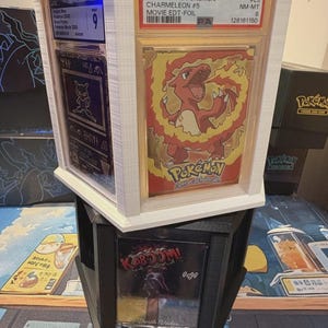 May include: A stack of three-dimensional display cases, each holding a collectible card. The top case features a graded 1999 Topps Pokemon Charmeleon card. The middle case has a Mew card. The bottom case displays a Darth Vader card with the word "KABOOM!"